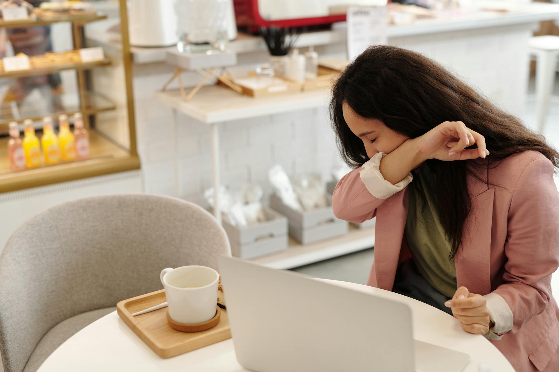 A woman seated at a table in a café, coughing into her elbow while sitting in front of a laptop, with a coffee cup on a tray 