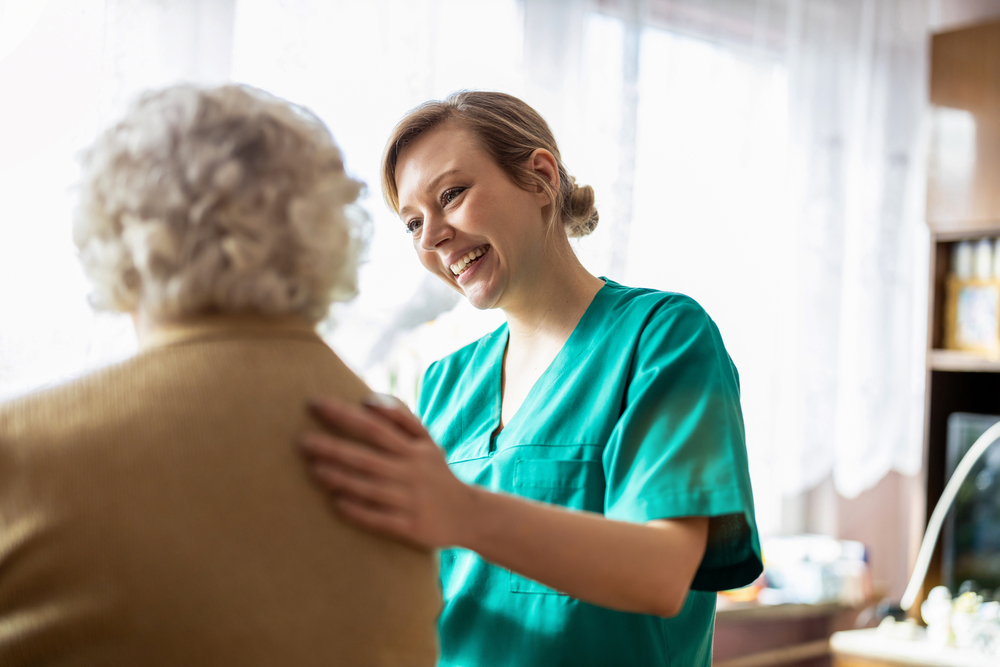 A smiling caregiver in green scrubs gently places her hand on the shoulder of an elderly person with gray hair.