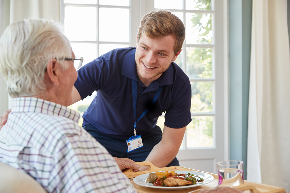 A support worker checking on an older man who is eating food.