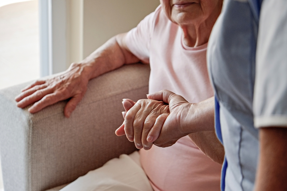 A close-up of a caregiver holding the hand of an elderly woman who is sitting on a chair.