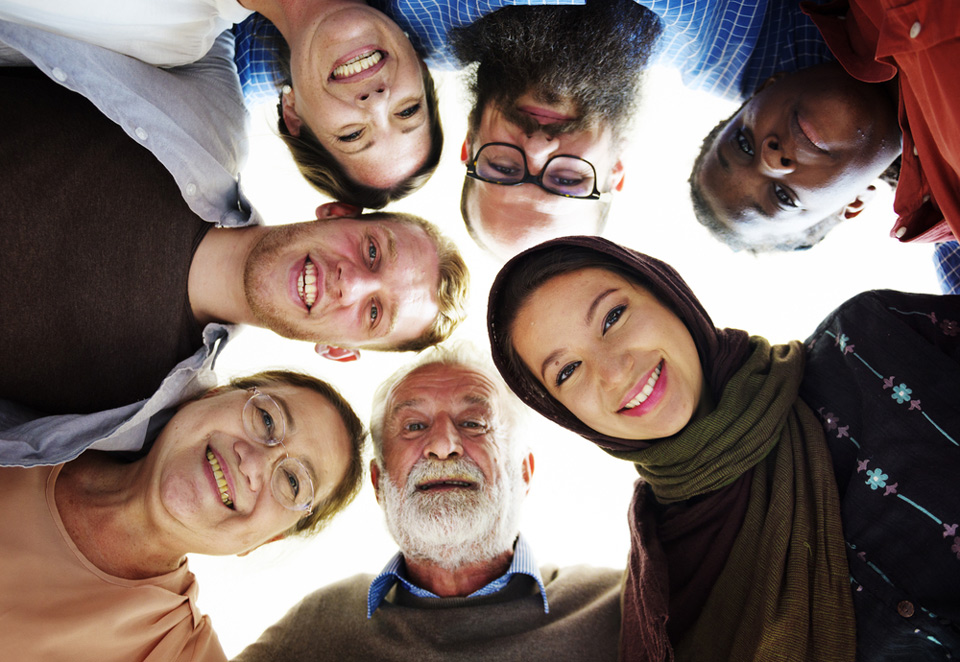 A diverse group of people of different ages, genders, and ethnicities stand in a circle, smiling down at the camera.