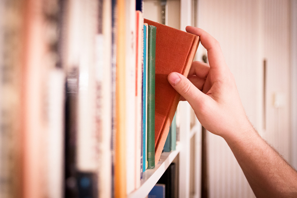 A hand reaching for a book on a shelf, selecting a hardcover book with an orange cover among a row of assorted books.
