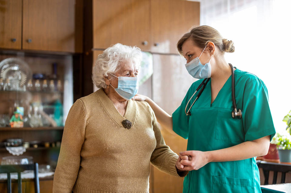 A nurse in green scrubs, wearing a face mask, comforting an elderly woman in a beige sweater, also wearing a mask.