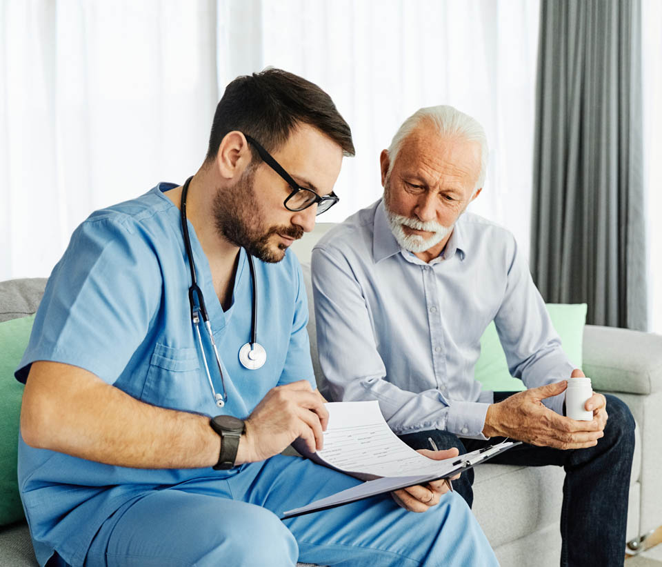 A doctor in a blue uniform reviews a clipboard with a seated elderly patient, holding a pill bottle, while discussing