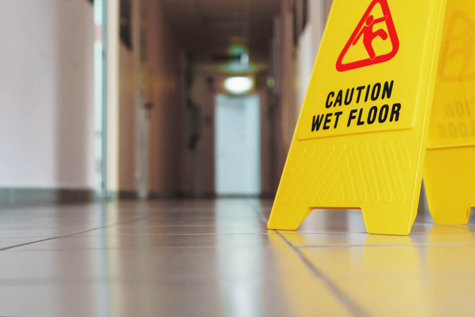 A yellow caution sign with the text 'Caution Wet Floor' stands on a tiled floor in a hallway. 