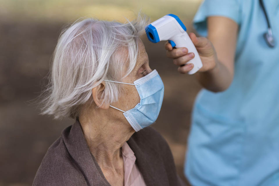 An elderly woman wearing a face mask gets her temperature checked by a nurse using a forehead thermometer.