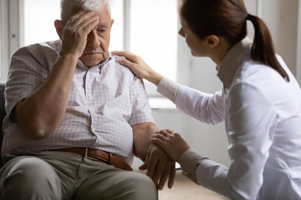 A female caregiver gently places a hand on the shoulder of an distressed elderly man sitting with a hand on his forehead