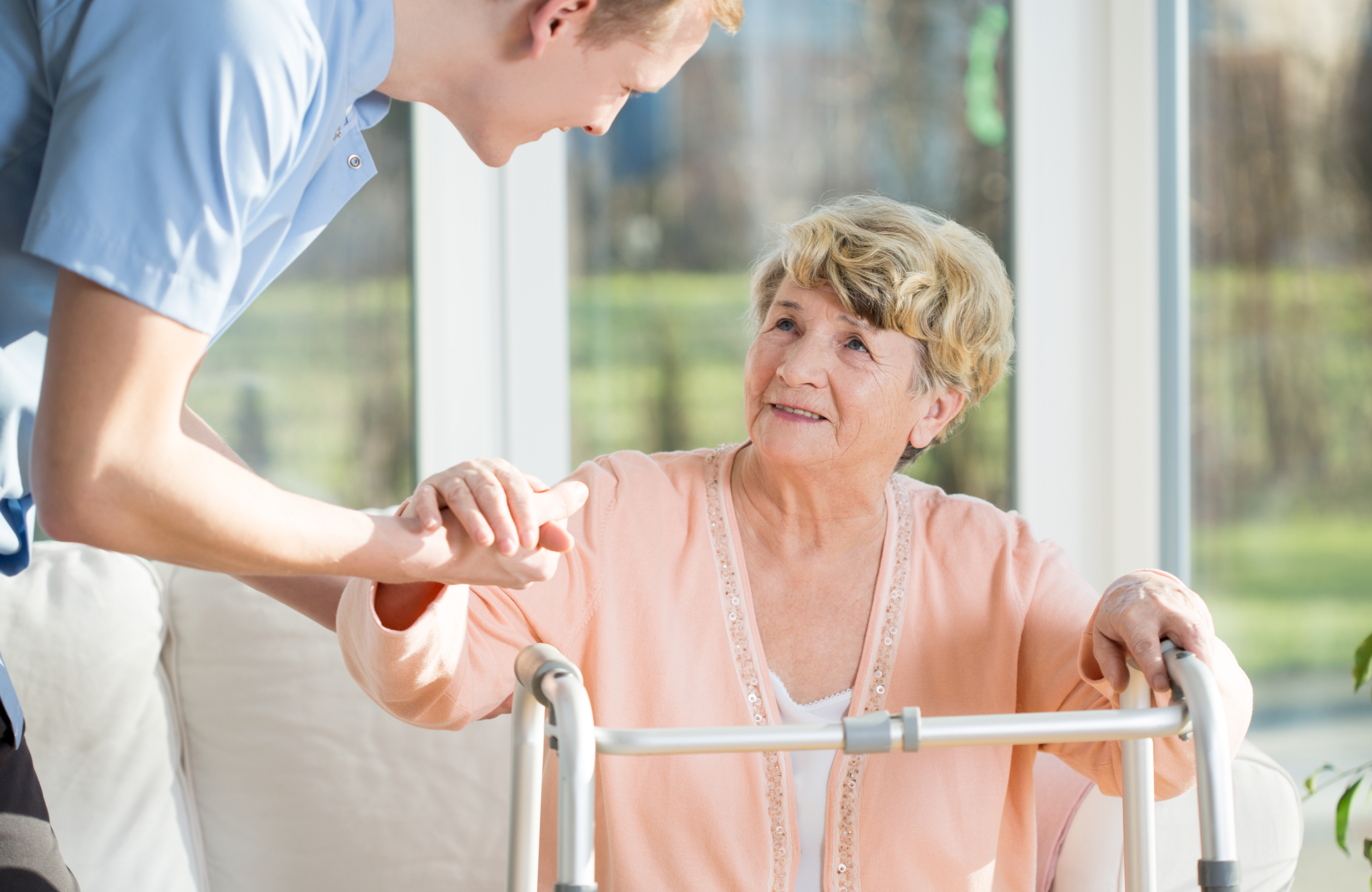An older woman is receiving assistance from a healthcare worker to stand from her chair.
